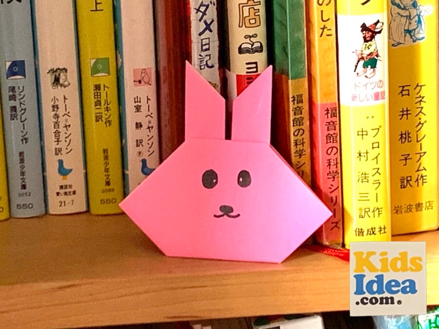 Origami "rabbit" placed in front of a book on the bookshelf ©︎2023 kids-idea.com